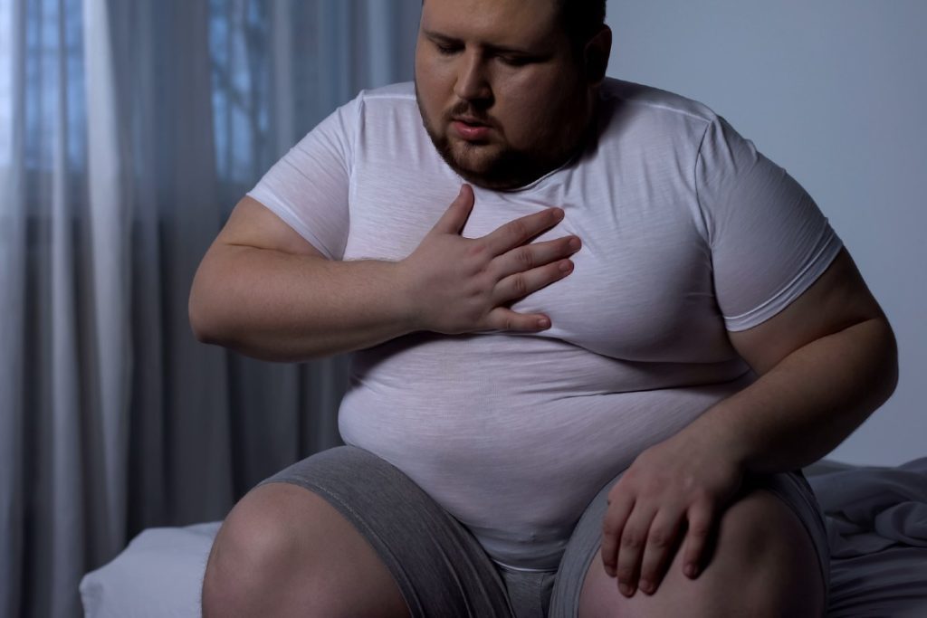 Übergewichtiger junger Mann hält sich, auf dem Bett sitzend, die Hand an die Brust
