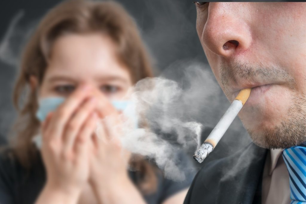 Frau unscharf im Hintergrund, die sich mit beiden Händen den Mund zuhält, während ein Mann im Vordergund eine Zigarette raucht