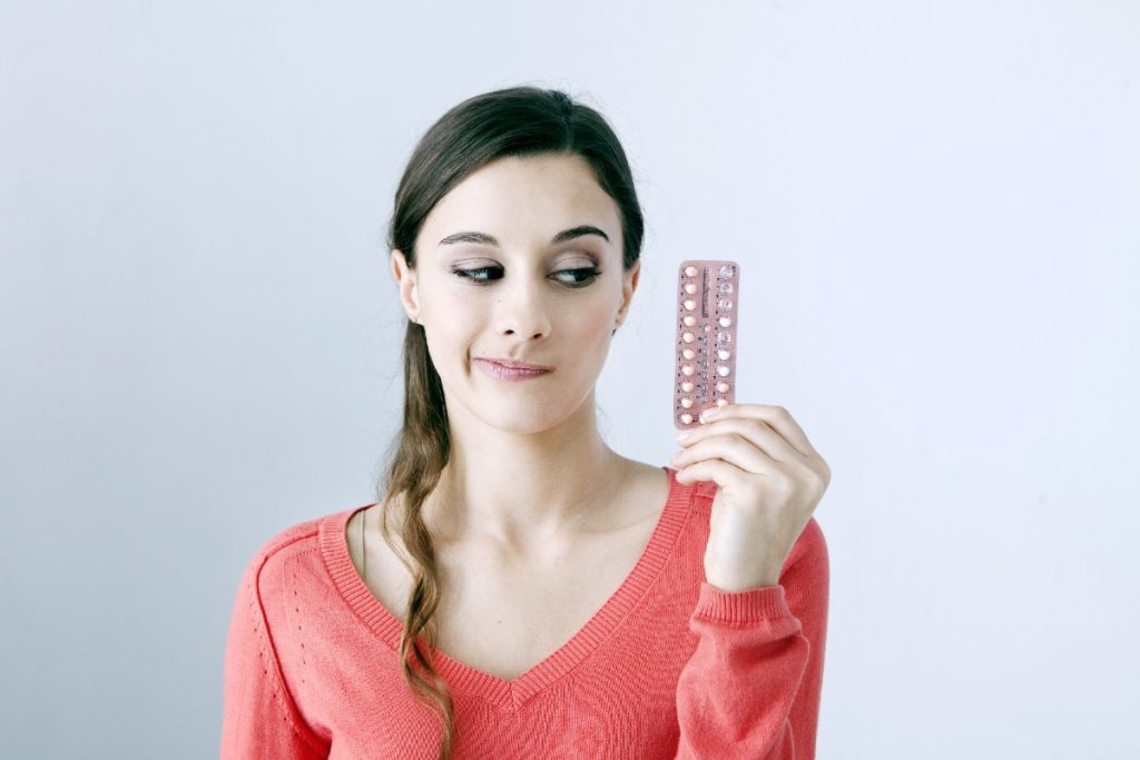 Junge Frau vor einheitlichem Hintergrund, die skeptisch auf eine Blisterpackung der Pille schaut