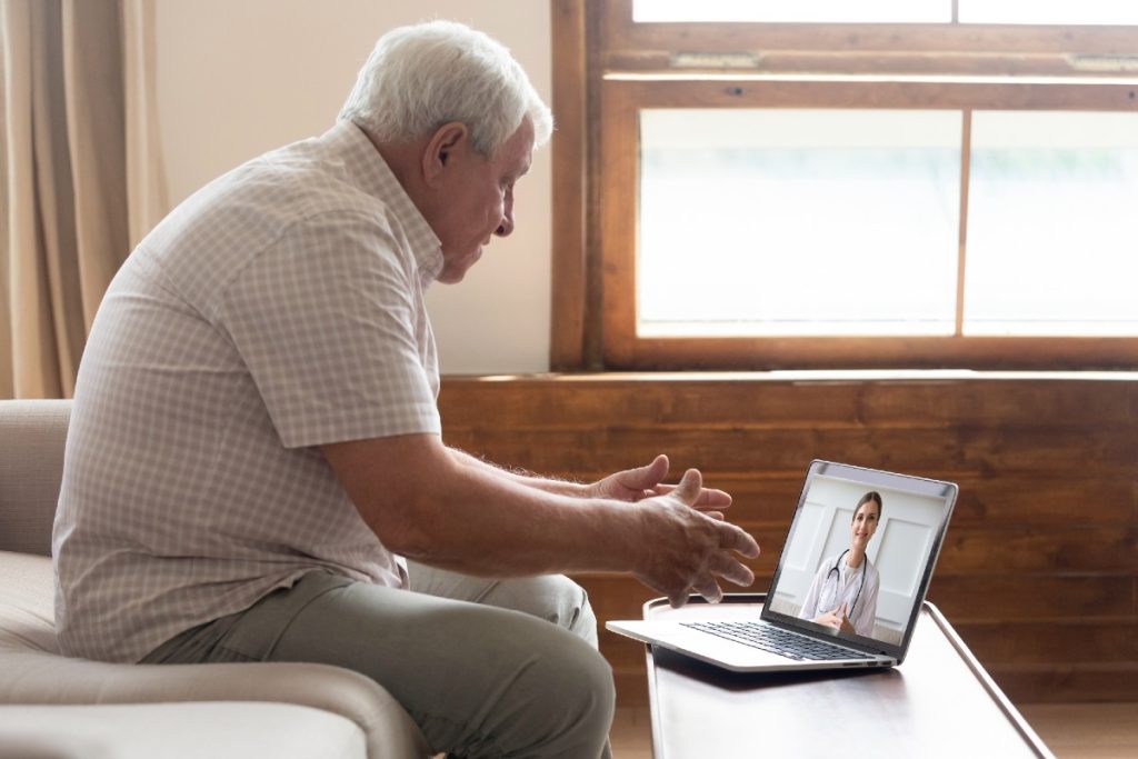 Älterer Mann am Wohnzimmertisch, der mit seiner Ärztin über Videosprechstunde am Laptop spricht
