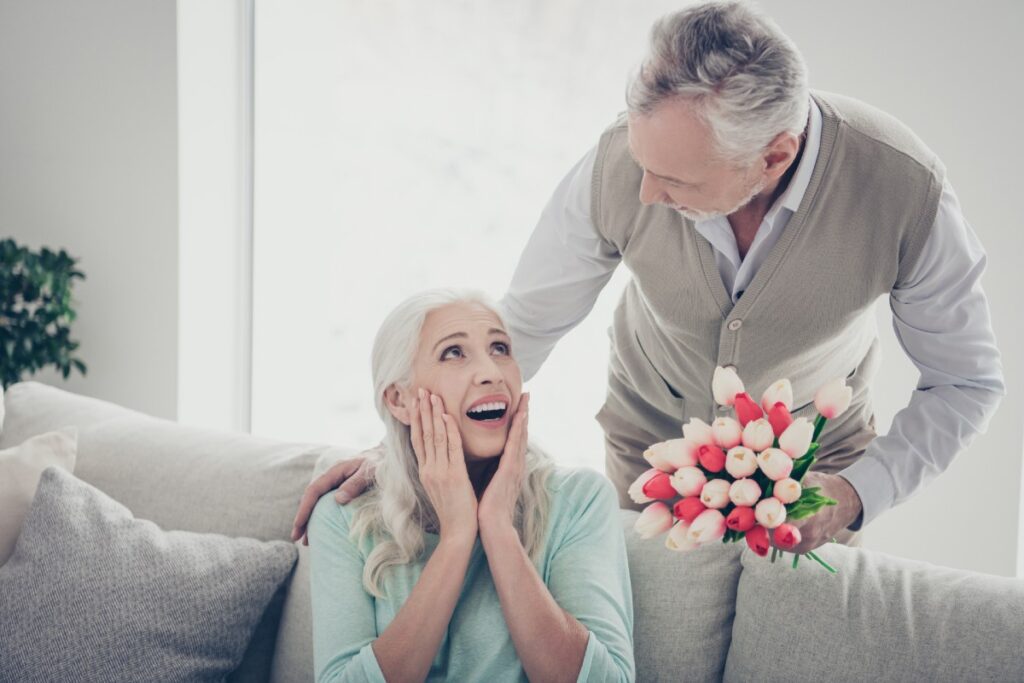 Älteres Paar, sie auf der Couch sitzend, er überreicht ihr einen Blumenstrauss