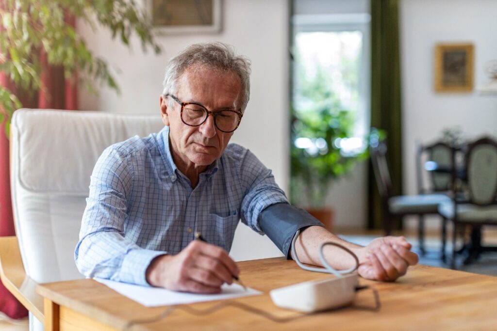 Älterer Mann am Tisch mit Blutdruckmessgerät, seine Werte auf ein Blatt Papier notierend