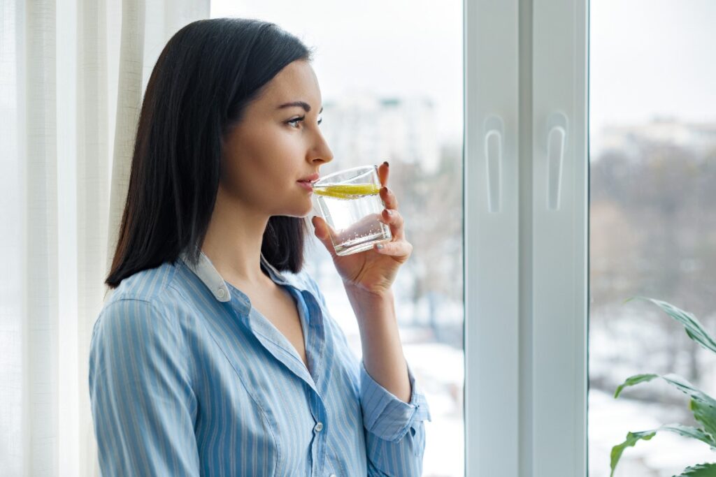 Junge Frau am Fenster, die ein Glas Wasser trinkt