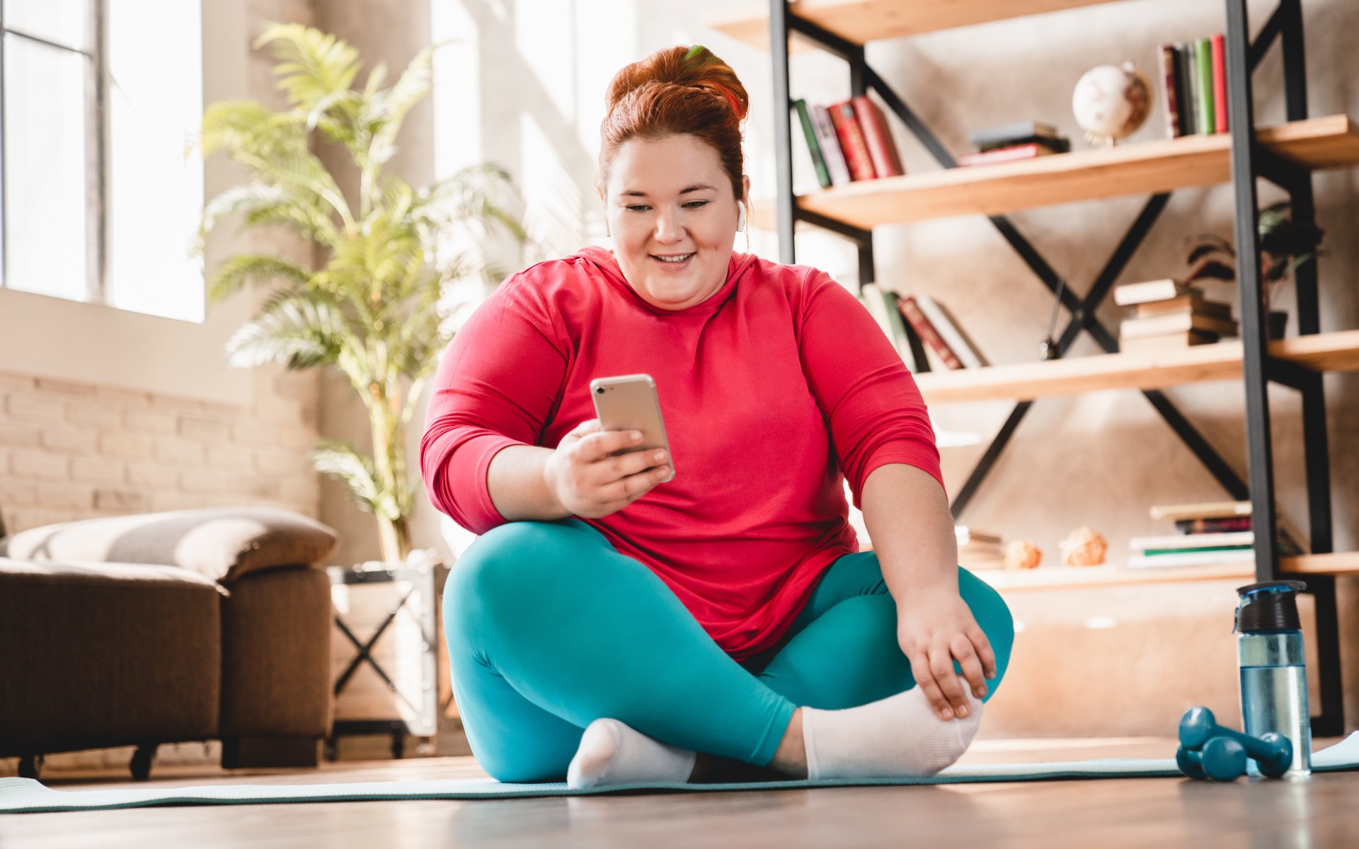 Übergewichtige junge Frau auf Yogamatte, lächelnd mit Handy
