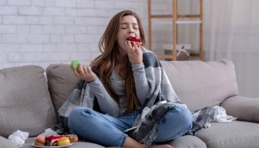 Junge Frau mit Decke über den Schultern auf der Couch sitzend, die Essen in sich hinein stopft