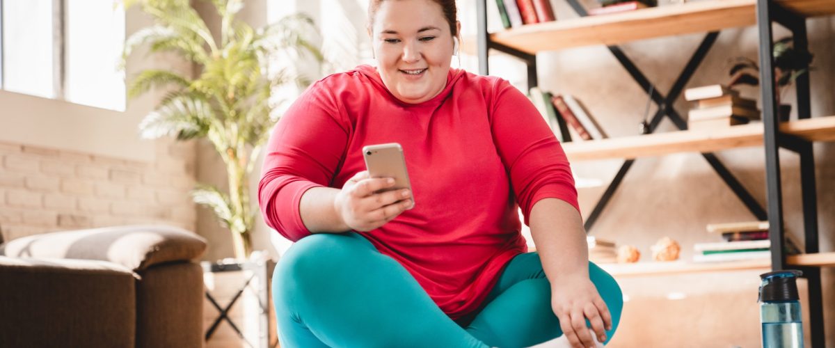 Übergewichtige junge Frau auf Yogamatte, lächelnd mit Handy