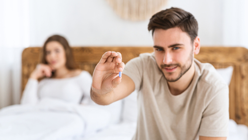 Pilule bleue tenue par un jeune homme souriant, représentant la santé masculine et la libido, avec une femme floue en arrière-plan dans une chambre moderne, gros plan et espace libre.