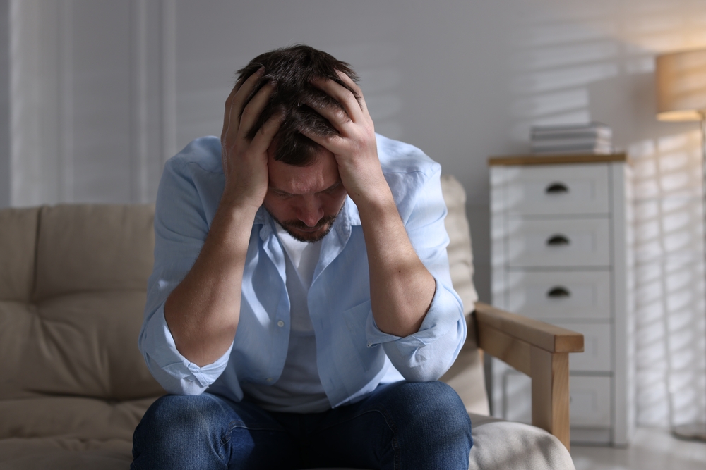 Homme adulte stressé et déprimé assis sur un canapé, tenant sa tête dans les mains, symbolisant la fatigue et la surcharge mentale masculine.