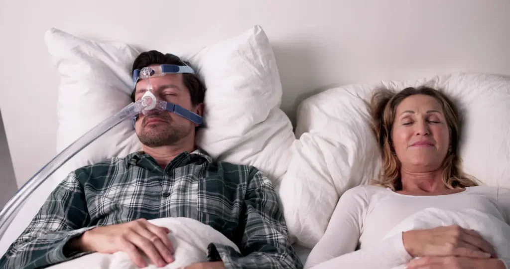 Pärchen im Bett, er mit CPAP, sie ist happy