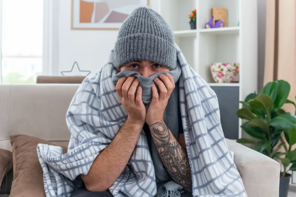 Ein schwacher, junger, kranker Mann mit Schal und Wintermütze sitzt auf dem Sofa im Wohnzimmer