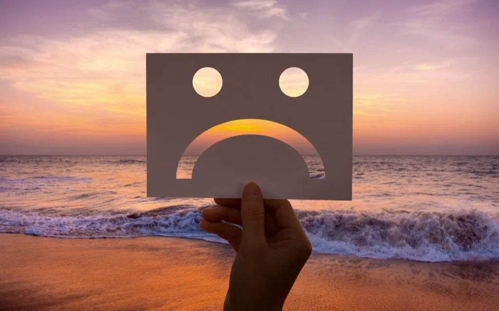 Ein trauriger Smiley in KArton ausgeschnitten vor einem Sonnenuntergang am Strand