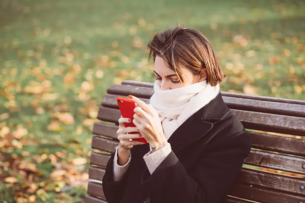 Frau mit hochgezogenem Schal sitzt im Herbst auf einer Parkbank und schaut auf ihr Smartphone.