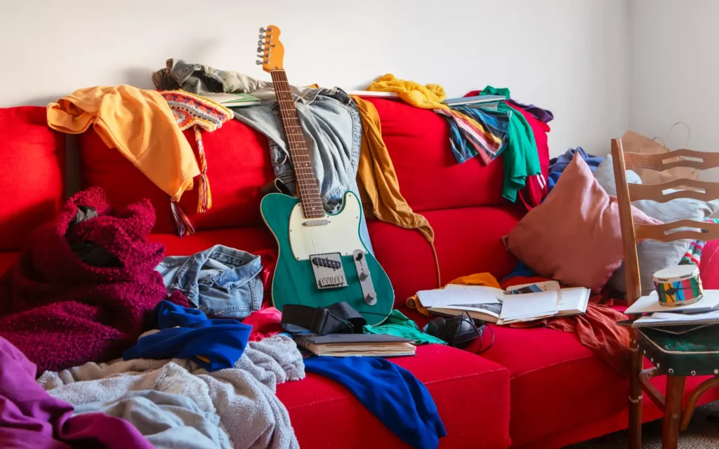 Unaufgeräumtes Wohnzimmer mit Kleidung, Büchern und Gegenständen auf einem roten Sofa.