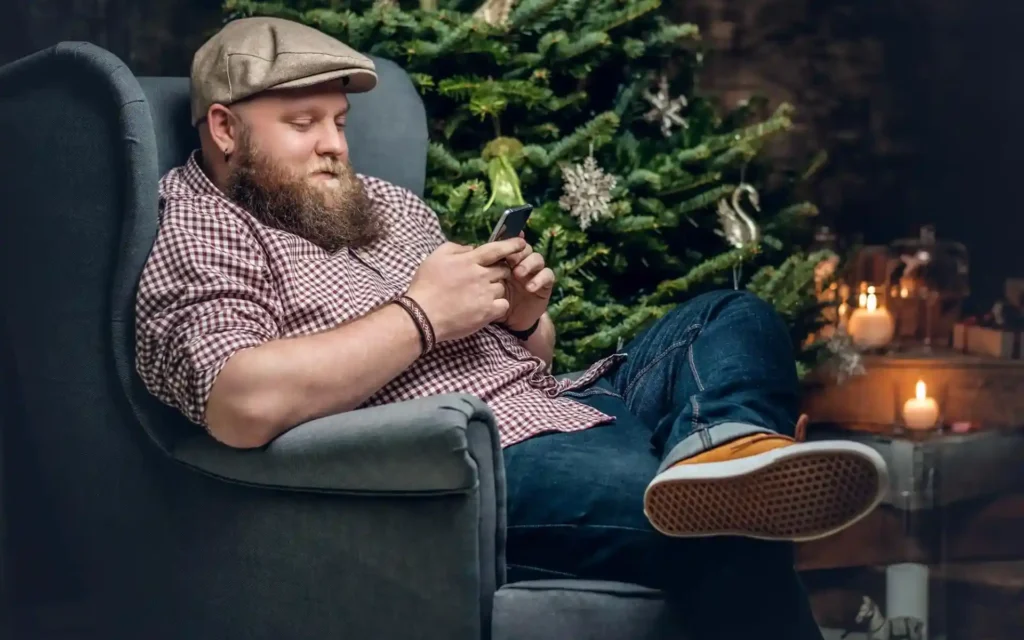 Dicker Mann mit Mütze sitzt vor einem Weihnachtsbaum in einem Sessel