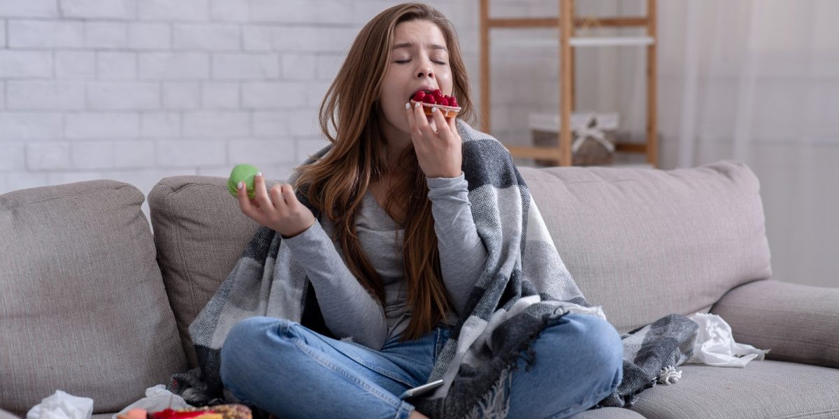 Junge Frau mit Decke über den Schultern auf der Couch sitzend, die Essen in sich hinein stopft