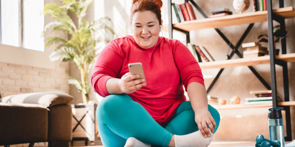 Übergewichtige junge Frau auf Yogamatte, lächelnd mit Handy