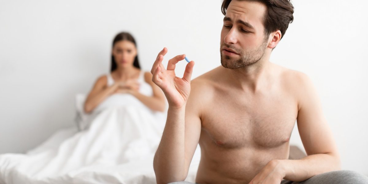 Junger Mann auf dem Bettrand sitzend, der eine Pille zwischen den Fingern hält, während ihn seine Partnerin im Hintergrund anschaut