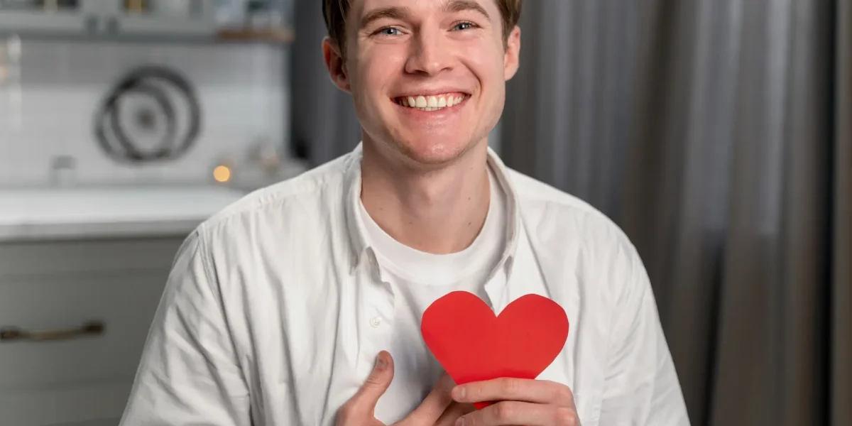Lächelnder Mann hält ein rotes Herz als Symbol für Herz-Kreislauf-Gesundheit und Cholesterinwerte.