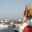 Junge Frau sitzt auf ihrem Koffer am Hafen und lächelt in die Kamera