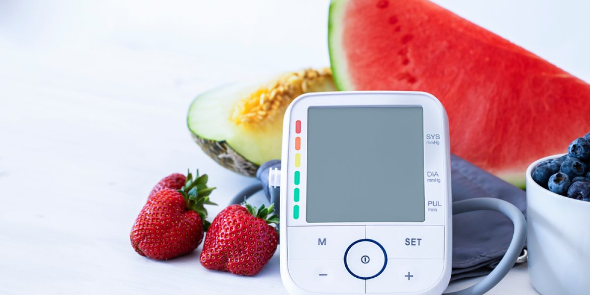 Écran de tensiomètre affichant une mesure de tension artérielle, posé sur une table en bois blanc à côté de fruits frais colorés