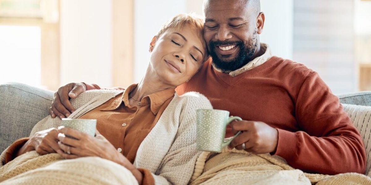 Un couple se détend sur leur canapé, chacun tenant une tasse de thé, les jambes couvertes d’une couverture, illustrant la détente et le bien-être hivernal.