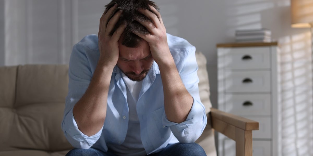 Homme adulte stressé et déprimé assis sur un canapé, tenant sa tête dans les mains, symbolisant la fatigue et la surcharge mentale masculine.